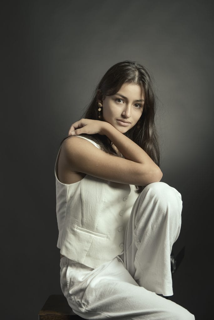 Portrait artistique d'une jeune femme. Pose assise, lumière douce et contrastée. Fond sombre. Ambiance studio cinema.