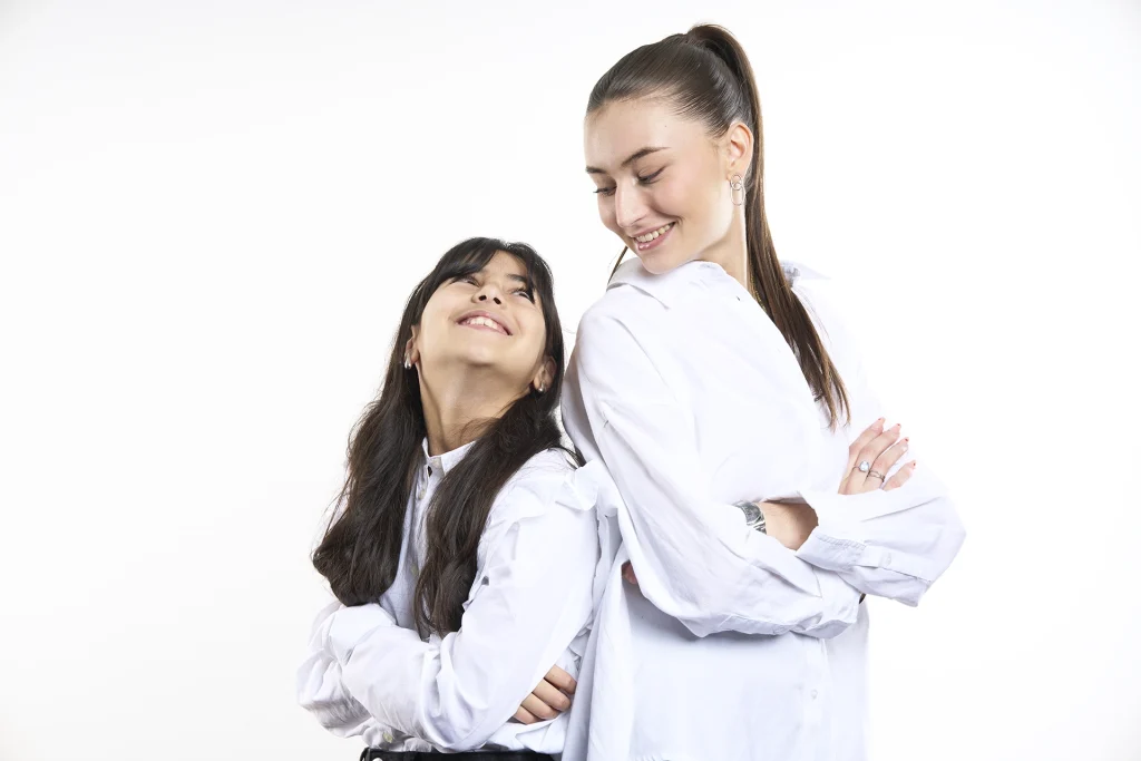 Portrait de 2 soeurs en couleur. Plan taille, lumière douce et contrastée. Visages souriants. Fond blanc