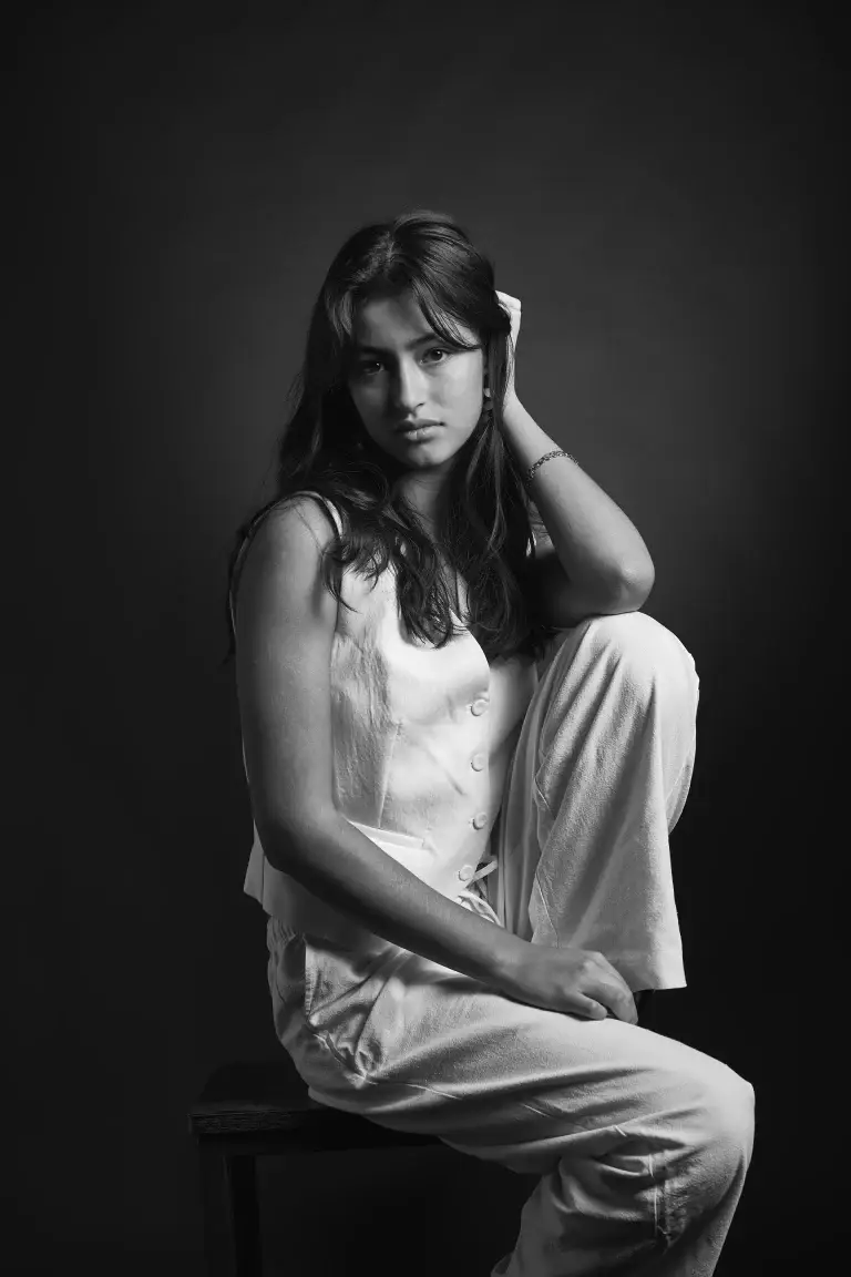 Portrait féminin artistique à tarbes. La modele est assise sur un tabouret. Ses vêtements clairs contrastent avec le fond sombre et la lumière zénitale.