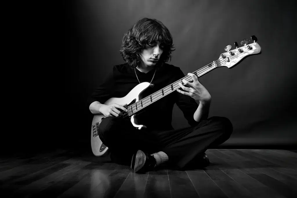 photo d'un musicien en noir et blanc. Le jeune homme est assis et joue de la basse. Décor et vêtements sombres.