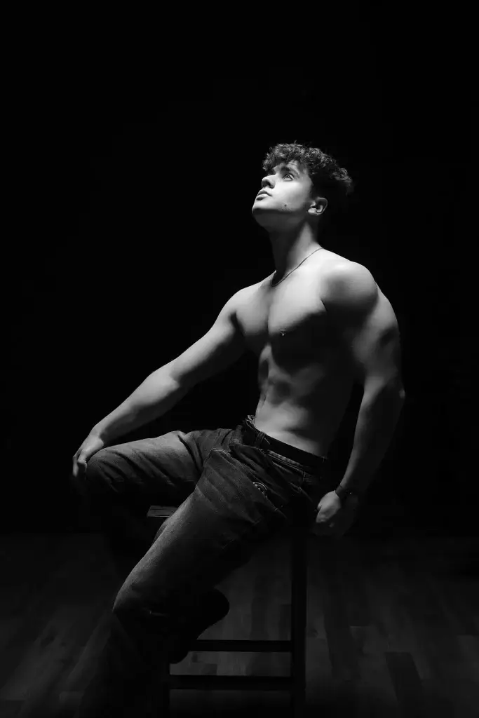 Portrait d'un jeune homme en noir et blanc et en clair obscur. La lumière en douche met en valeur sa musculature et son attitude générale. Pose assise cadrage de pieds.