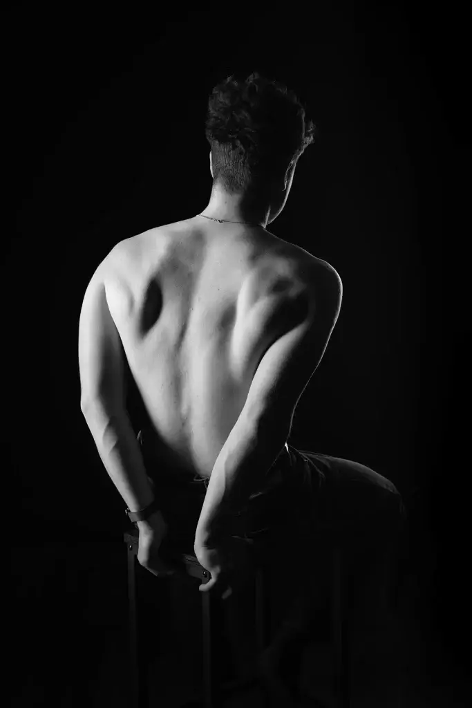 Portrait en noir et blanc et en clair obscur d'un homme assis de dos. La lumière met en valeur la musculature de son dos.