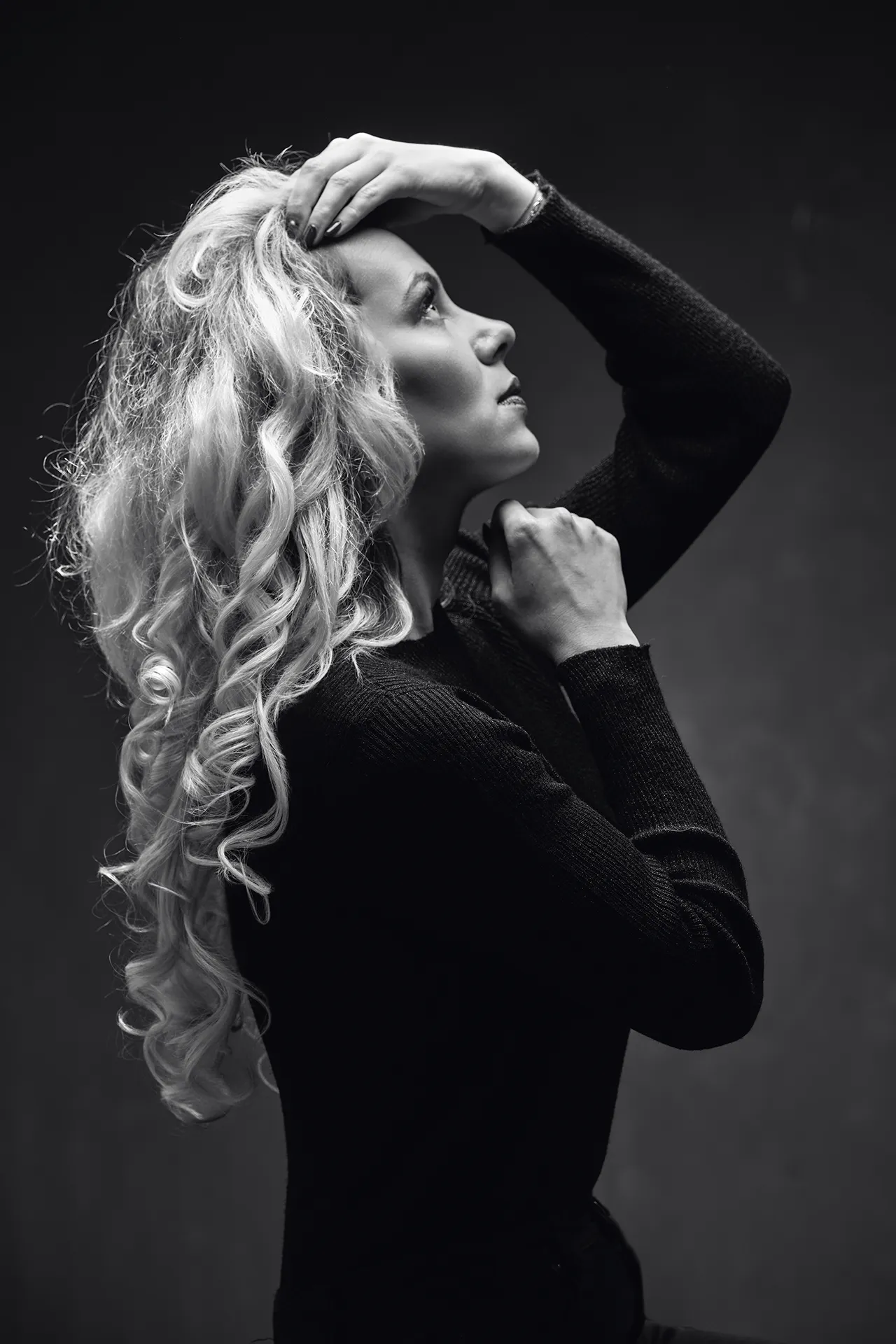 Portrait en noir et blanc d'une jeune femme blonde assise. La photo est prise de profil dans un plan italien. La lumière en douche éclaire son visage. Sa silhouette sombre se détache du fond qui est éclairé.
