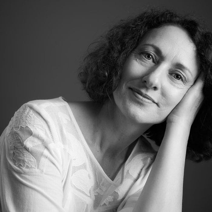 Portrait de studio en noir et blanc : femme aux cheveux bouclés, chemise blanche, joue posée sur la main.