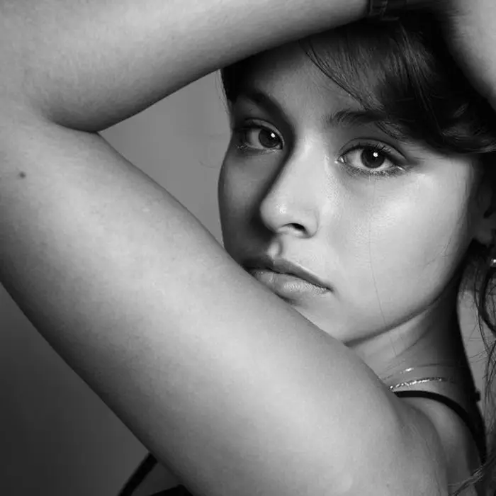 Portrait de studio en noir et blanc, cadrage serré : jeune femme, bras replié au-dessus de la tête, regard face à l'objectif.
