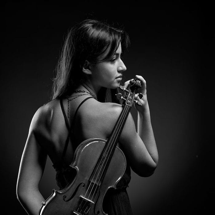 Portrait de studio en noir et blanc : violoniste vue de dos, violon posé sur l'épaule, fond sombre.